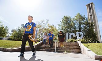 Low angle of a student, wearing a UCR shirt, walking with his skateboard and other students, also wearing UCR branded merch, walking down the nearby staircase.